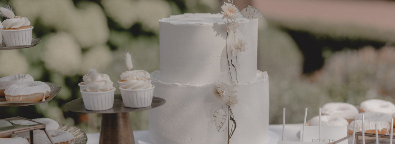 Zweistöckige weiße Hochzeitstorte mit Trockenblumen auf einem Sweet Table mit Cupcakes und Donuts